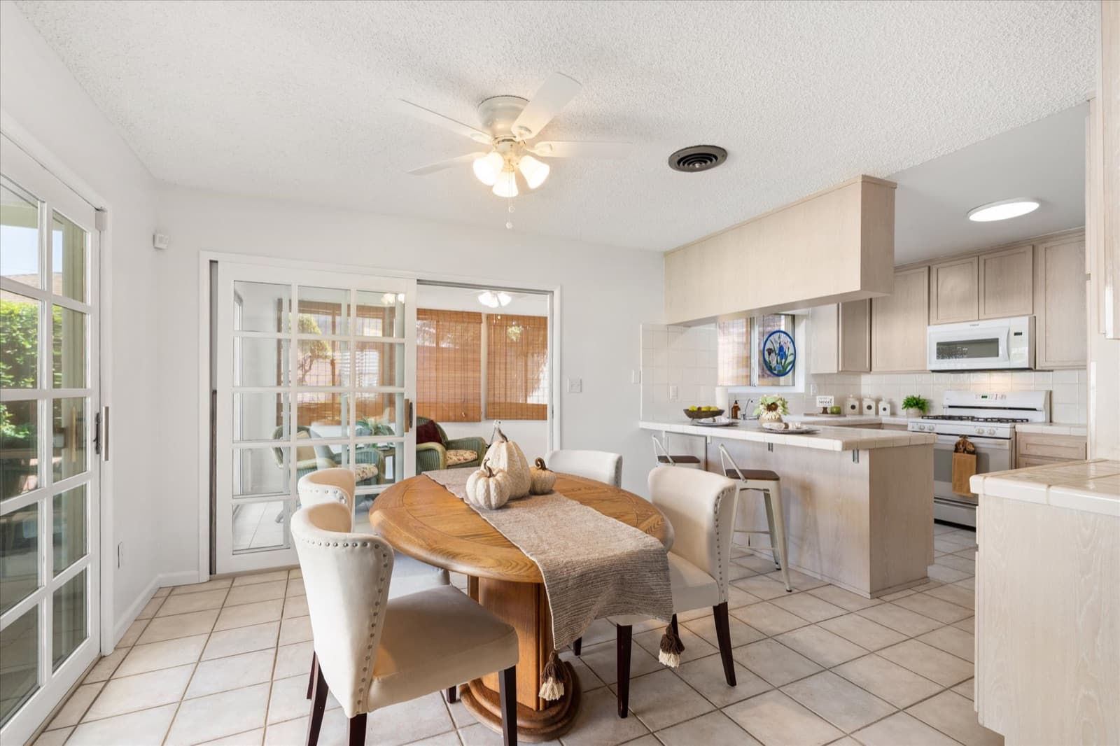 Dining area looking through to the kitchen, with a wood table on tile flooring, the breakfast bar, and French doors to the enclosed sunroom