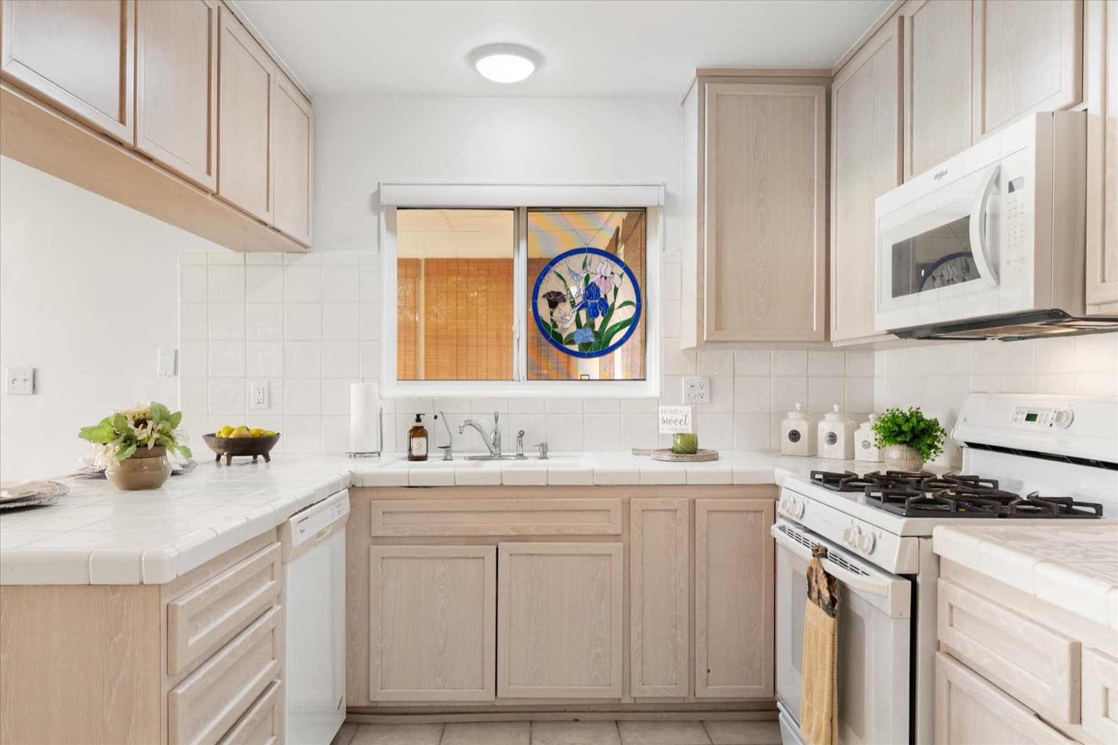 Kitchen with whitewashed wood cabinets, tile countertops, a white gas range, and a stained-glass iris medallion in the window above the sink