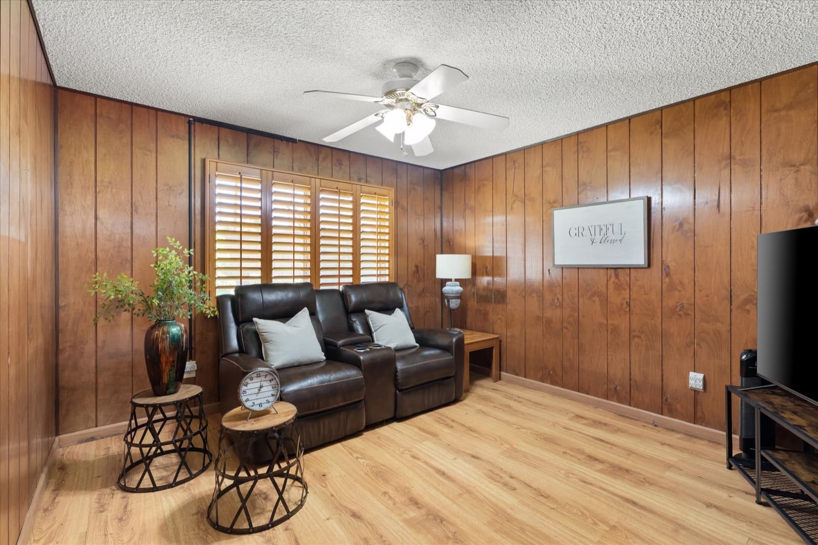 Wood-paneled den with a leather reclining loveseat, plantation shutters, and light wood flooring