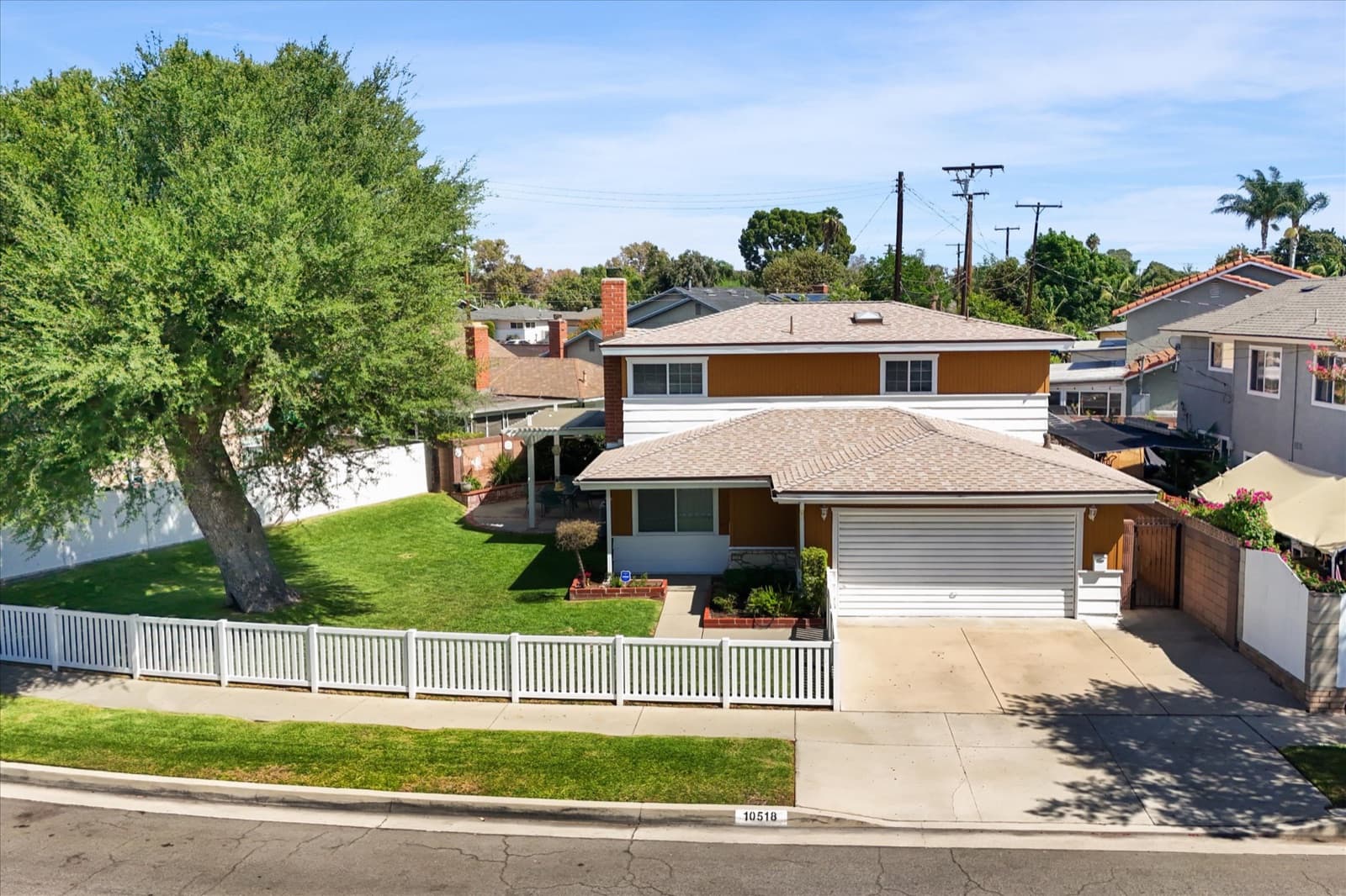 Low aerial view of the front of the property showing the driveway, white picket fence, front lawn, and neighboring rooftops