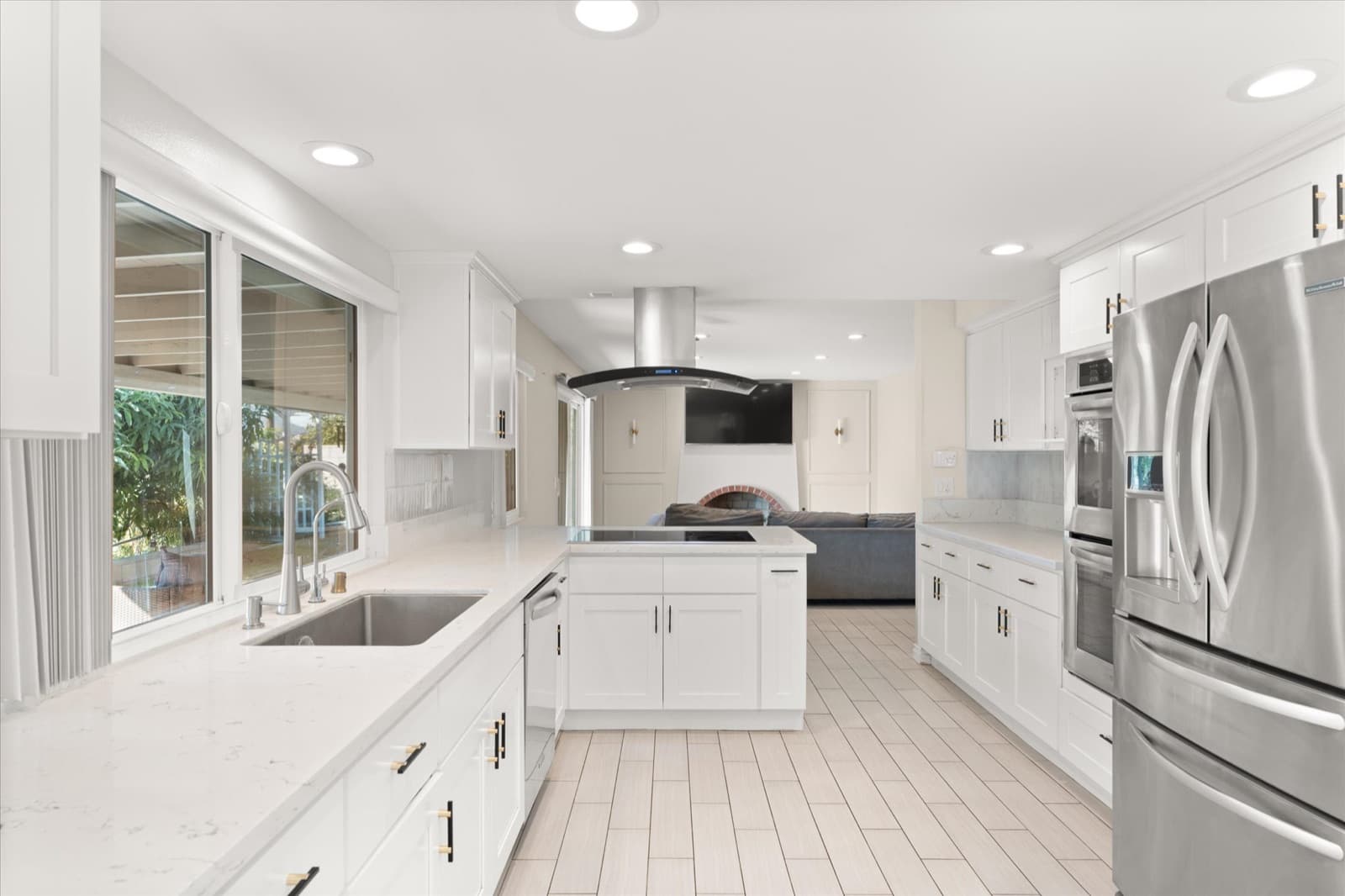 White kitchen with an island cooktop and range hood, stainless steel appliances, and windows over the sink looking into the backyard