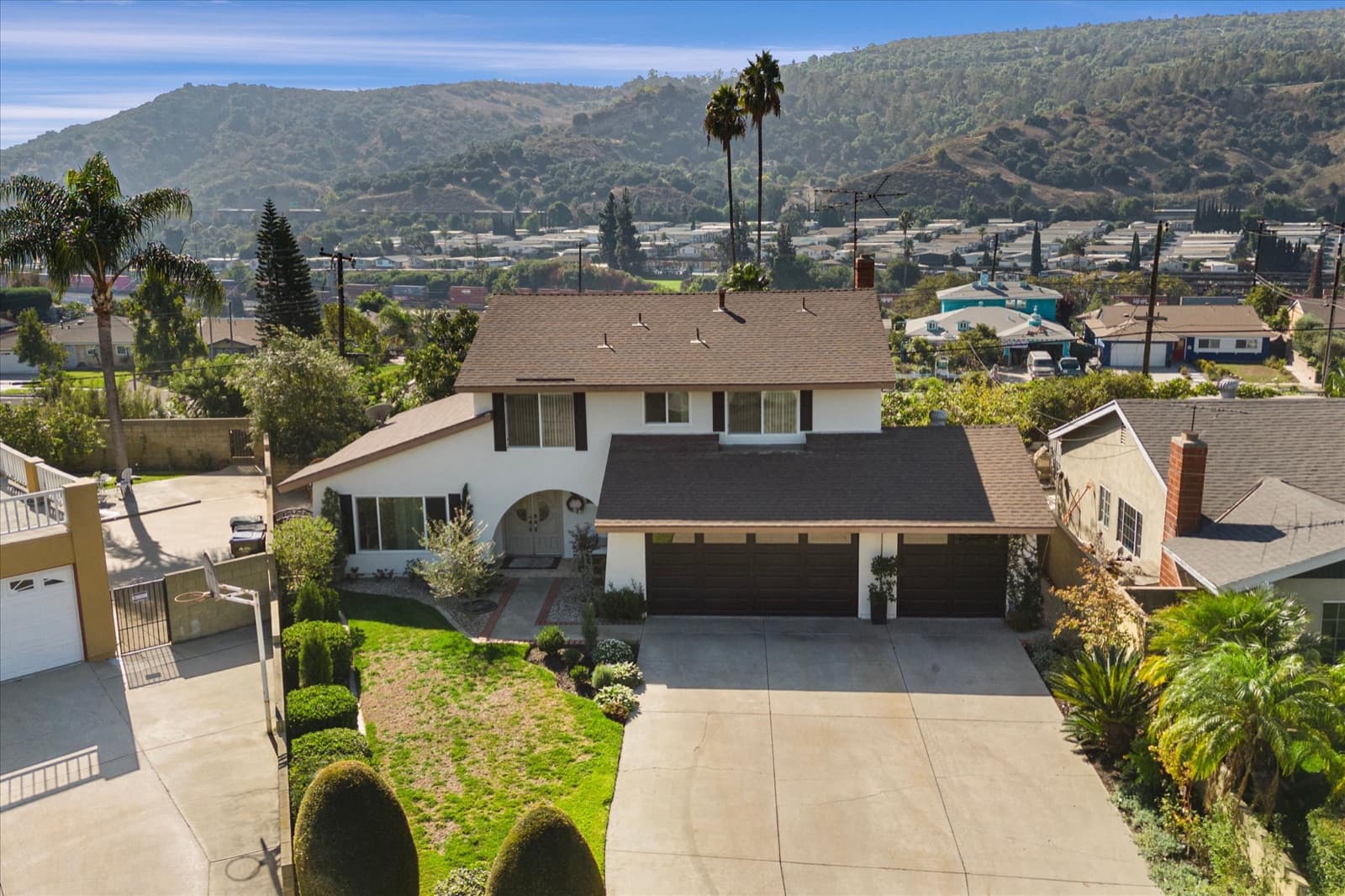 Low aerial view of the home, its driveway and front lawn, with the Whittier foothills behind